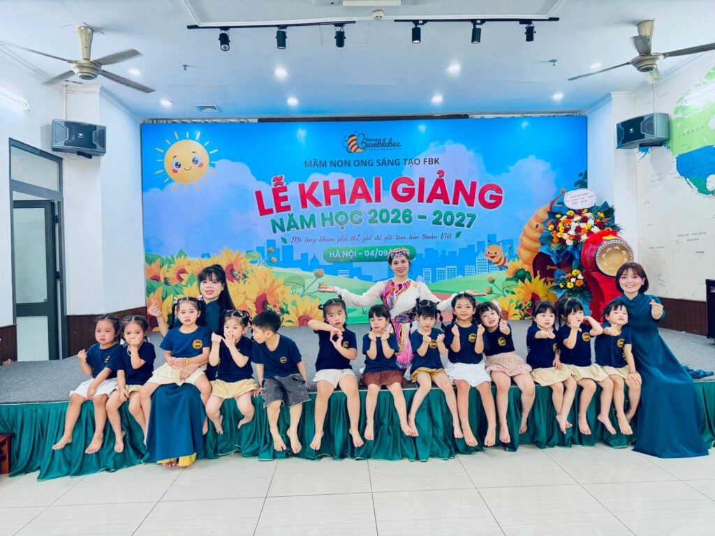 Các con FBK mặc áo đồng phục ngồi trên mép sân khấu cùng cô giáo