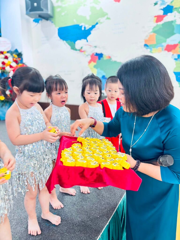 Cô giáo bưng khay quà hình con ong cho các con chọn, phía sau là bản đồ thế giới